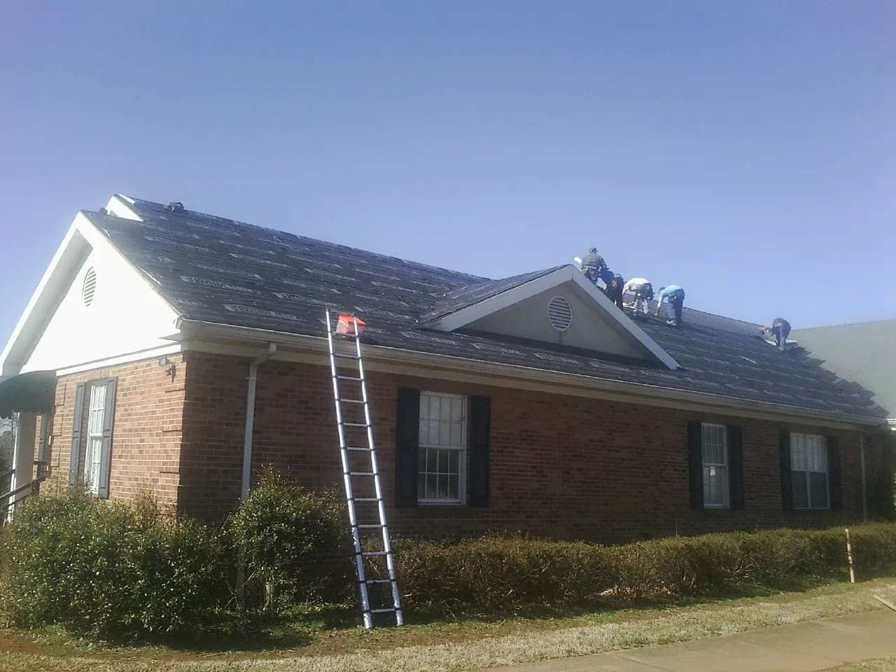 Crew tearing shingles off a brick ranch roof with a ladder set — Emergency Roof Repair work by our crew in Riverside